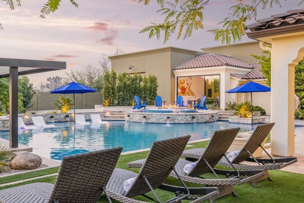 Luxury backyard with resort-style pool, fire pit, lounge chairs, blue umbrellas, and modern home at dusk