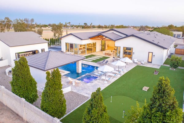 Modern white house with dark roof, blue swimming pool, patio seating, manicured lawn, and putting green, aerial view