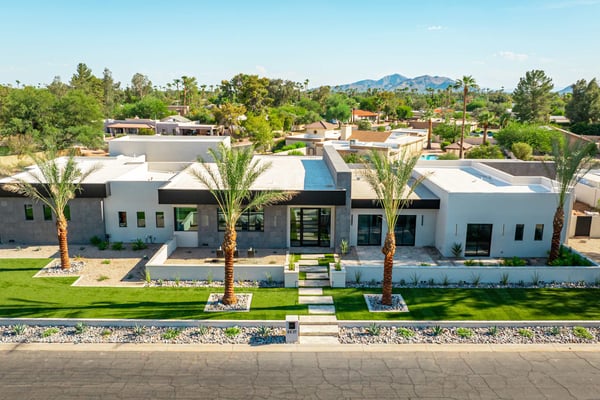 Modern white and concrete home with flat roof, palm trees, and manicured lawn in desert neighborhood with mountains in background