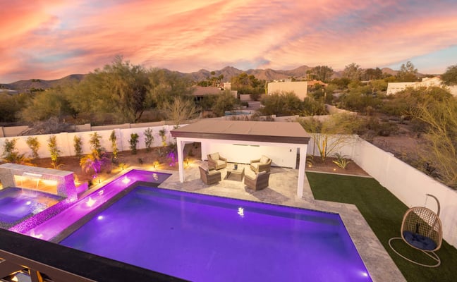 Backyard with illuminated purple pool, spa, lounge seating, and desert mountains at sunset