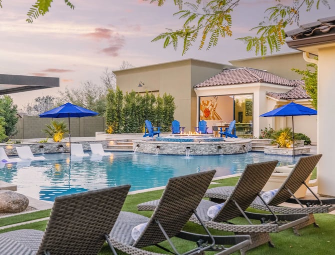 Luxury backyard with resort-style pool, fire pit, lounge chairs, blue umbrellas, and modern home at dusk
